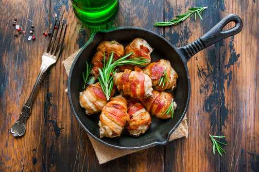 Oven roasted bacon wrapped chicken drumsticks in a black baking pan on the wooden rustic table, top view.